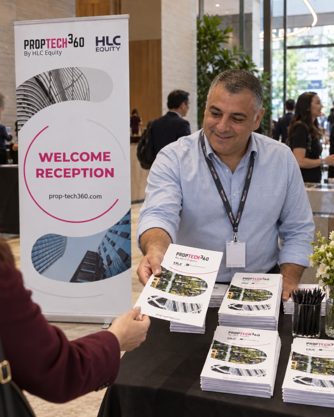 A receptionist distributing PropTech360 handouts beside the reception banner.