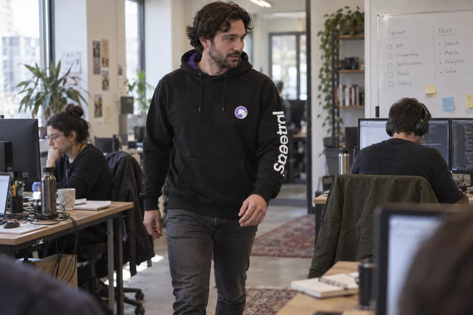 An engineer walking between shared office desks wearing the original black Spectral hoodie.